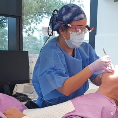 A person in a mask, safety glasses, gloves and hair net shines a light into the mouth of a prone man in pink clothing.